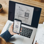 Faktura z kodem QR Osoba przy laptopie skanuje telefonem kod QR wyświetlony na fakturze elektronicznej, mając obok papierową wersję faktury z KSeF. Temat: KSeF, faktury elektroniczne, kody QR i ryzyko pustej faktury.