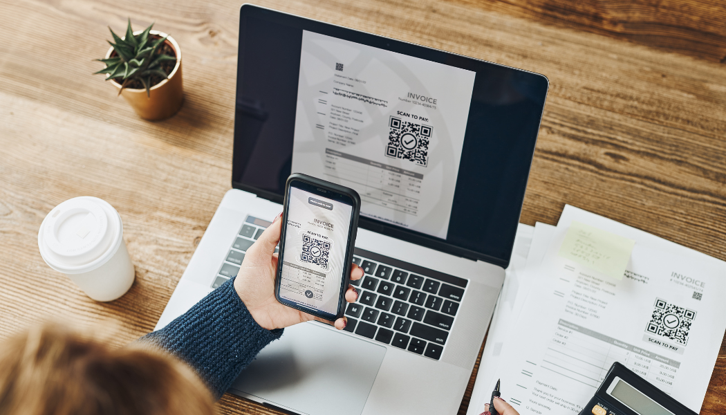 Osoba przy laptopie skanuje telefonem kod QR wyświetlony na fakturze elektronicznej, mając obok papierową wersję faktury z KSeF. Temat: KSeF, faktury elektroniczne, kody QR i ryzyko pustej faktury.